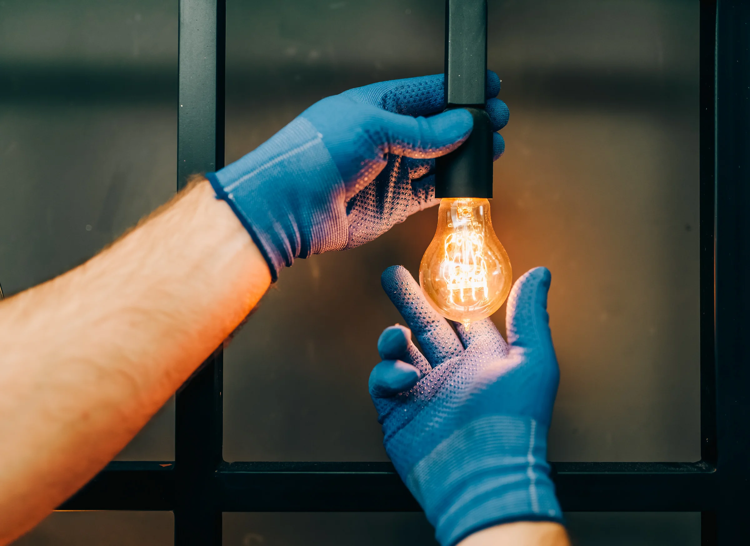 Hände mit blauen Schutzhandschuhen schrauben eine leuchtende Edison-Glühbirne in eine Fassung vor Metallrahmen