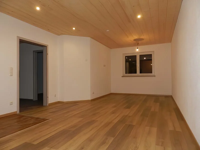 Leerstehender Raum mit hellem Parkettboden, Holzdecke mit Einbauleuchten, weiße Wände, Fenster und offene Tür
