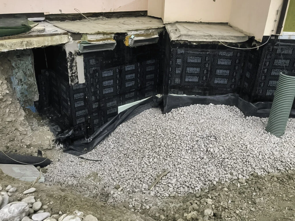 Ausgegrabene Kelleraußenwand an Ecke mit befestigter schwarzer Noppenbahn auf Fundament und Schotterbett am Fuß