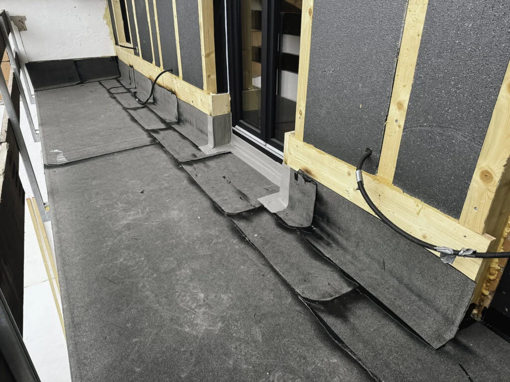 Schwarze Flüssigkunststoff-Abdichtung an Wandfuß und Flachdachanschlüssen, nahtlose Schichten um Tür-/Fensteröffnung.