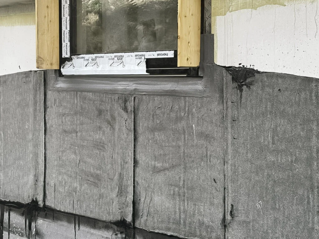 Grau aufgetragene Flüssigkunststoff-Schicht auf Mauerwerk unter Fensterlaibung mit sichtbarer Holzunterkonstruktion