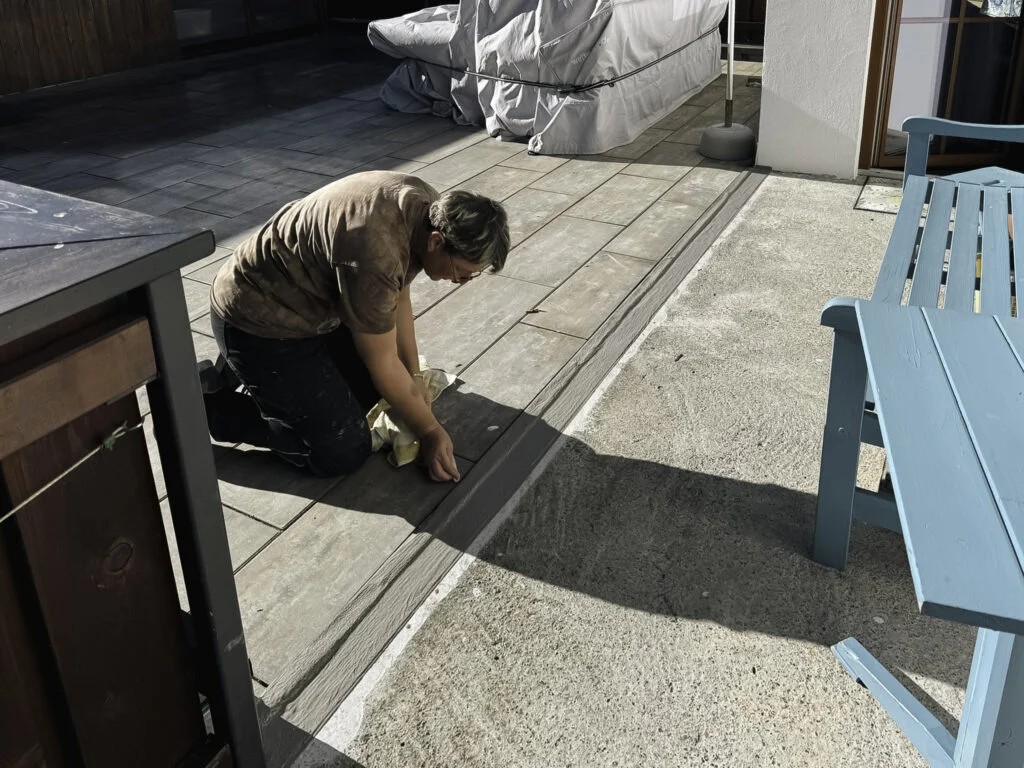 Person kniet auf Betonterrasse und bearbeitet Fugen mit Spachtel; Geländer links, Tür und Arbeitsmaterial im Hintergrund.