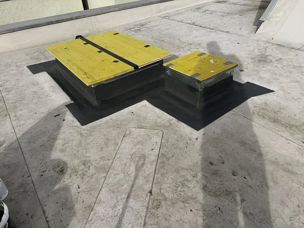 Flachdach mit zwei gelben Wartungsluken auf mit Flüssigkunststoff abgedichteten Sockeln, graue Dachfläche