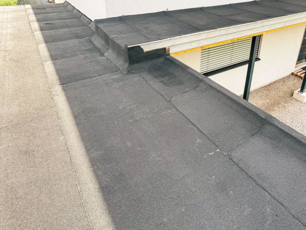 Flachdach mit schwarzer Bitumenabdichtung, sichtbaren Nähten, Metallabdeckung an Brüstung und seitlichem Dachrand