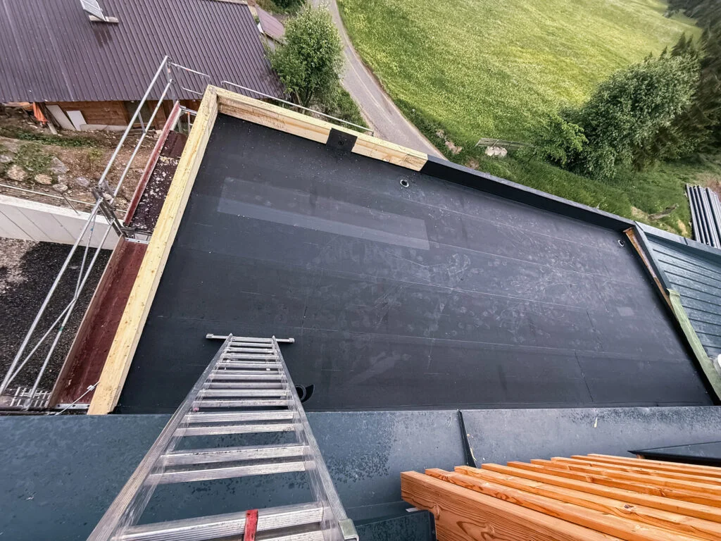 Flachdach mit dunkelmatten Abdichtungsbahn, aufgehender Holzkastenrand, Leiter und Gerüst an der Dachkante, umgeben von Gras.