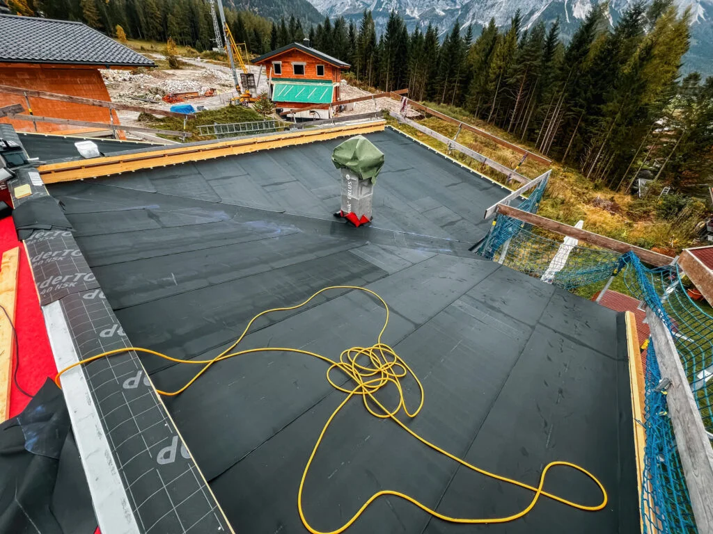 Flachdach mit Abdichtungsbahnen, Kran-Transportkorb, Kabel/Schläuche und Personen bei Dachabdichtungsarbeiten, umgeben von Wa