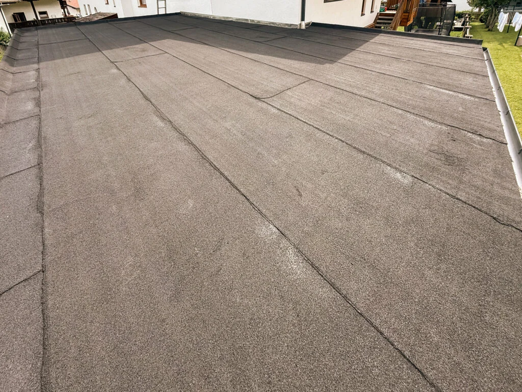 Flachdach mit grau-braunen Abdichtungsbahnen, sichtbaren Stößen und Nahtfugen, niedrige Dachkante und Gebäudewand.