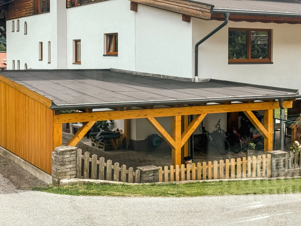 Holzcarport mit dunklem Flachdach, darunter Holzstapel und Lattenzaun; angrenzendes weißes Haus mit Schrägdach.