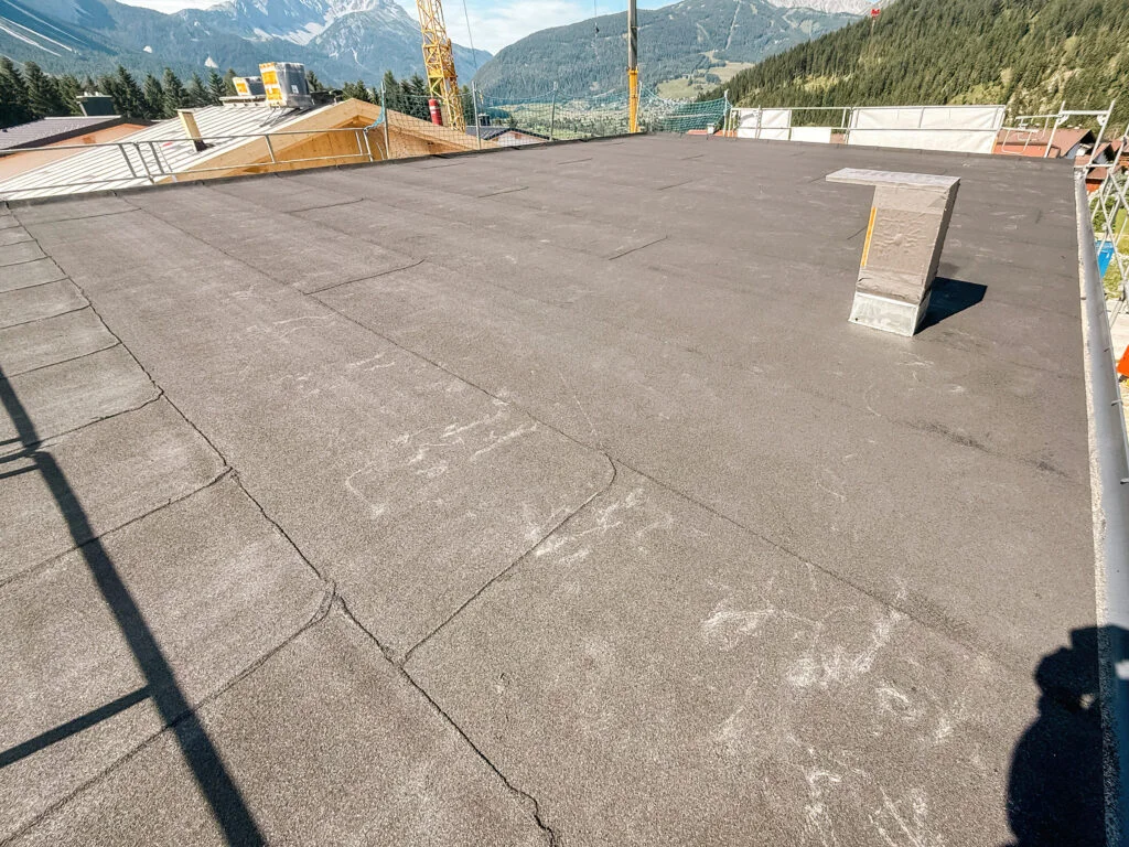 Flachdach mit bituminöser Dachbahn, Randabschlüsse und Entlüftungsrohr; Berglandschaft am Horizont.
