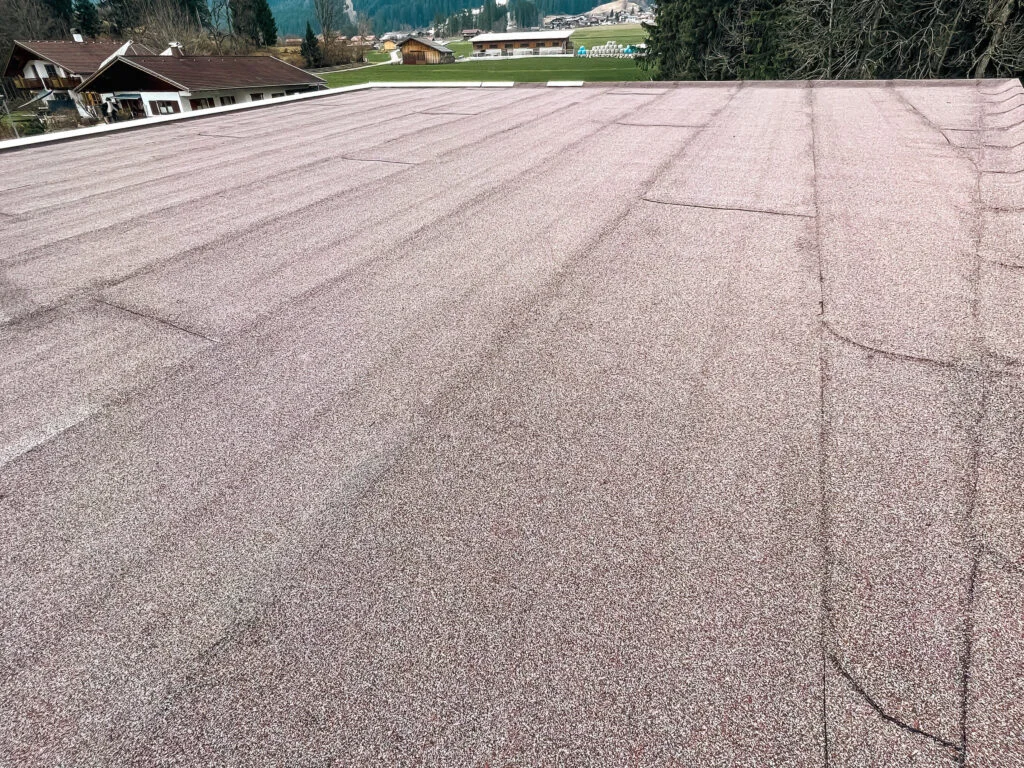 Flachdach mit hellbraunen Mineralgranulatbahnen, sichtbaren Stoßnähten und Randabschlüssen; im Hintergrund Bäume und Häuser.
