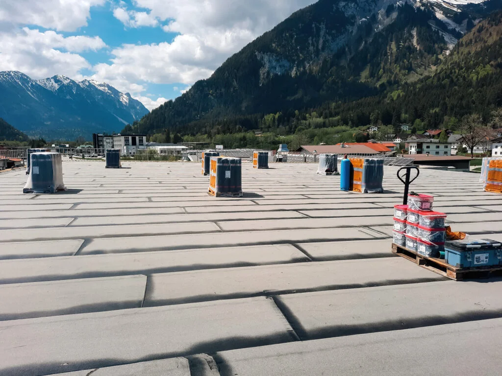 Flachdach mit aufgerollten Abdichtungsbahnen, Werkzeug und Material auf der Dachfläche; Bergkette im Hintergrund