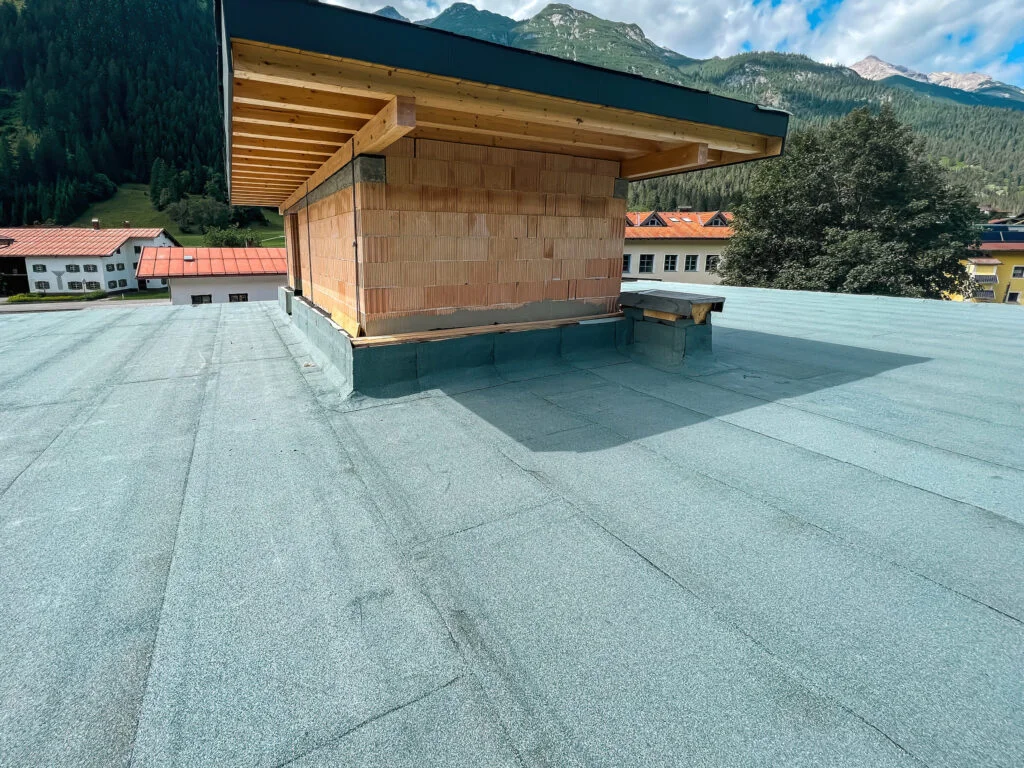 Flachdach mit Bitumenbahn, hölzerner Dachaufsatz/Schornstein und Bergen im Hintergrund