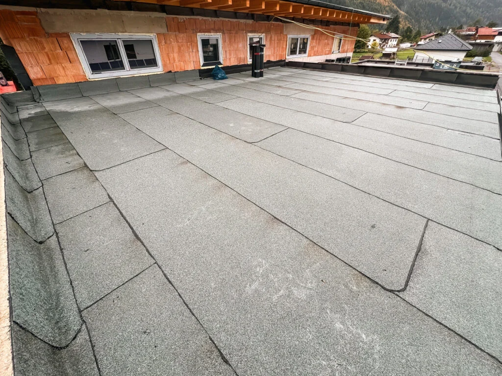 Flachdach mit überlappenden Dichtungsbahnen und sichtbaren Nähten, angrenzende orange Fassade mit Fenstern und Türen