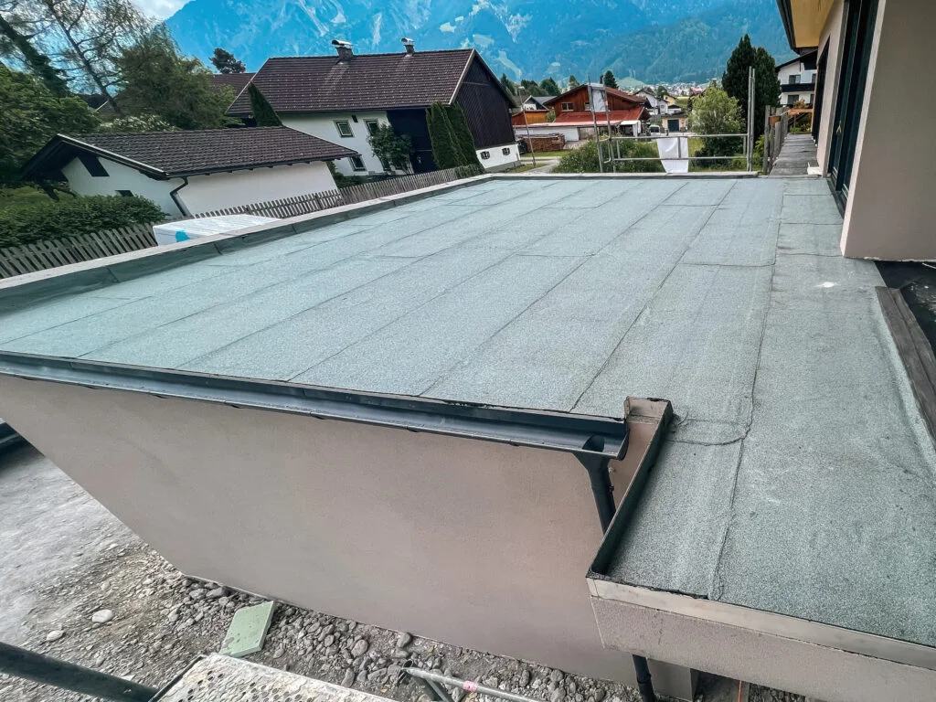 Hellgraue, mineralisch belegte Dachbahn auf Flachdach mit umlaufender Brüstung und Metallabdeckung; Wohnhäuser im Hintergrund