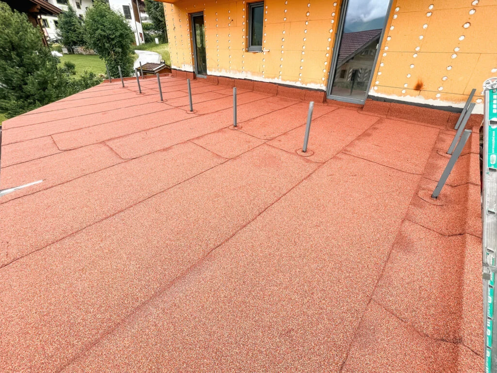 Flachdach mit rotbrauner Schweißbahndecke, mehrere Entlüftungsrohre, orange Außenwand mit Tür und Fenster