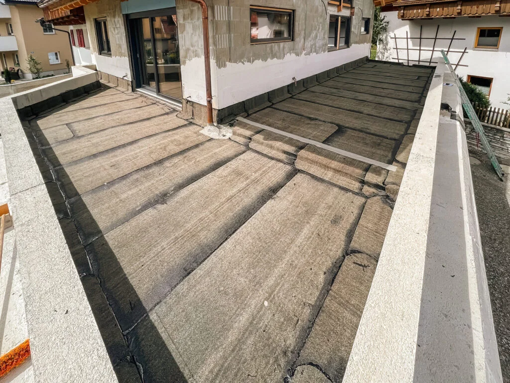 Flachdach mit aufgebrachten Bitumenbahnen und überlappenden Nähten, angrenzende Hauswand mit Tür und Fenster.