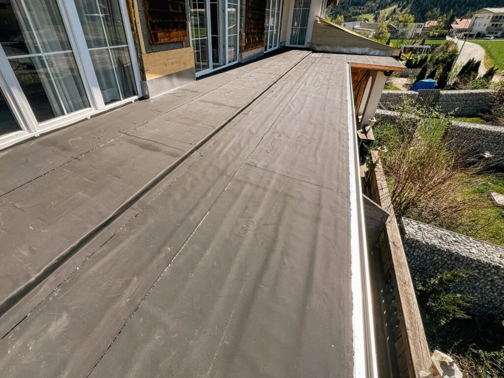 Flachdach mit aufgeklebter Abdichtungsbahn, Metallrand und Rinne an einer Terrasse mit großen Glasschiebetüren