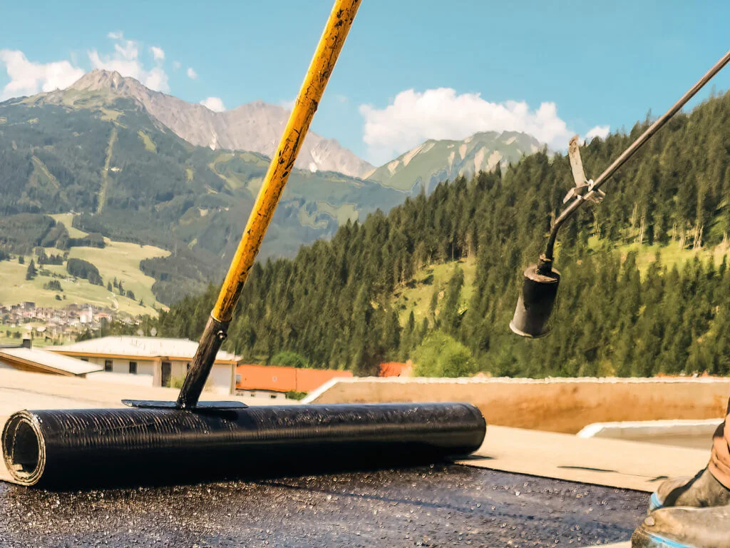 Walze und Schaufel tragen schwarze Abdichtungsmasse auf Flachdach auf; im Hintergrund Berge, Wald und Dächer.