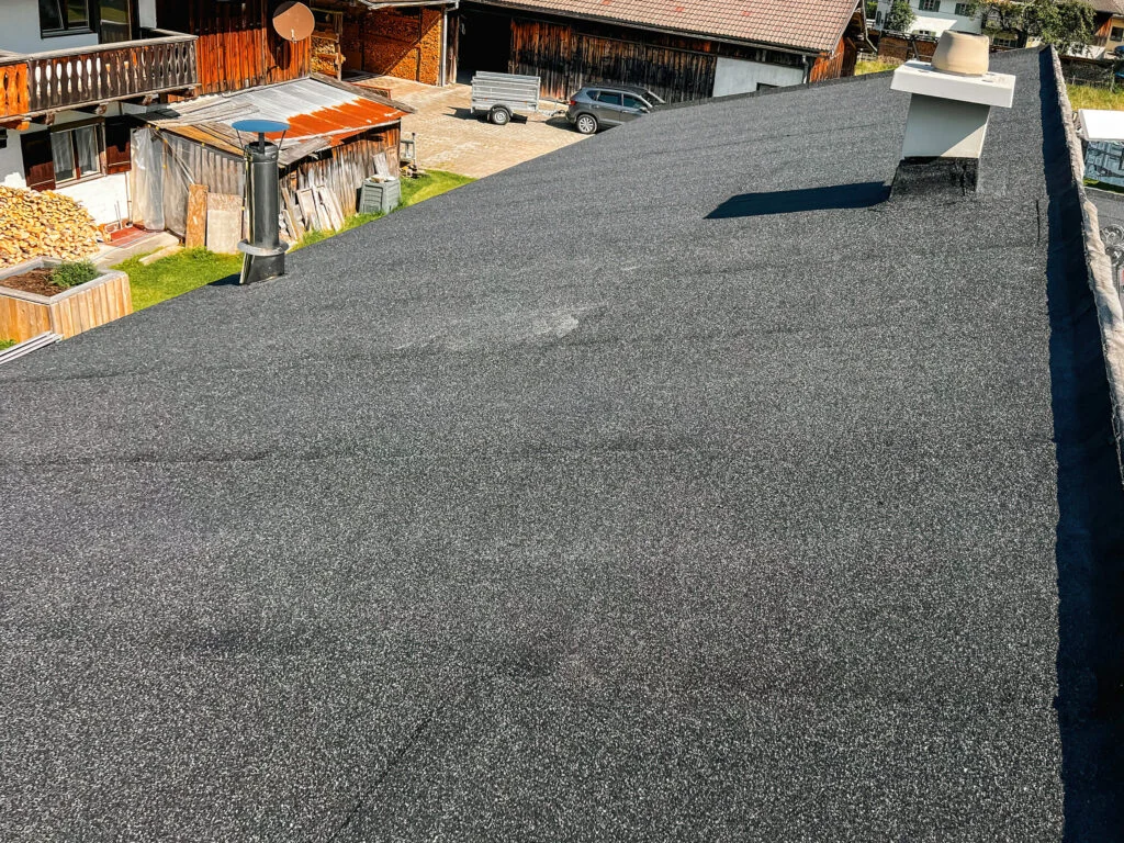 Flachdach mit dunkler, körniger Abdichtungsoberfläche und leicht erhöhtem Dachrand; Wohnhäuser im Hintergrund.