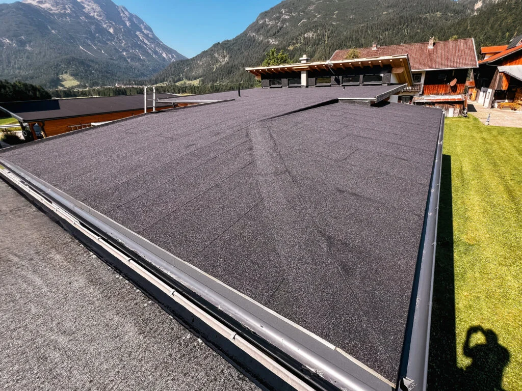 Flachdach mit dunkler Schüttung/Decklage, Attika und Regenrinne; im Hintergrund Berge und Häuser.