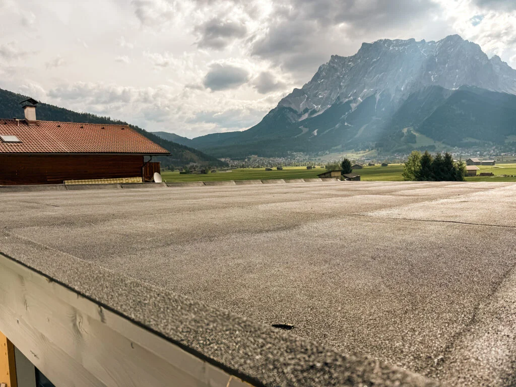 Flachdach mit körniger Abdichtungsoberfläche und niedriger Attika, im Hintergrund Tal, Gebäude und bewölkte Berge.