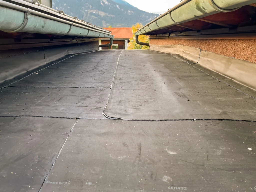 Flachdach mit dunkelgrauer Abdichtungsbahn zwischen niedrigen Brüstungen; Rinnen links und rechts; Bergkulisse im Hintergrund