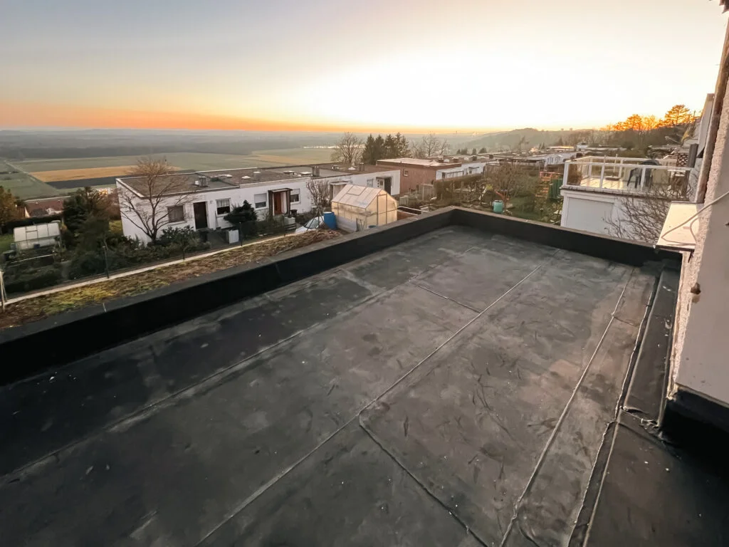 Flache Terrasse mit dunkler Abdichtungsbahn, niedrige Brüstung und Geländerpfosten; Blick auf Ort und Berge bei Abendlicht.