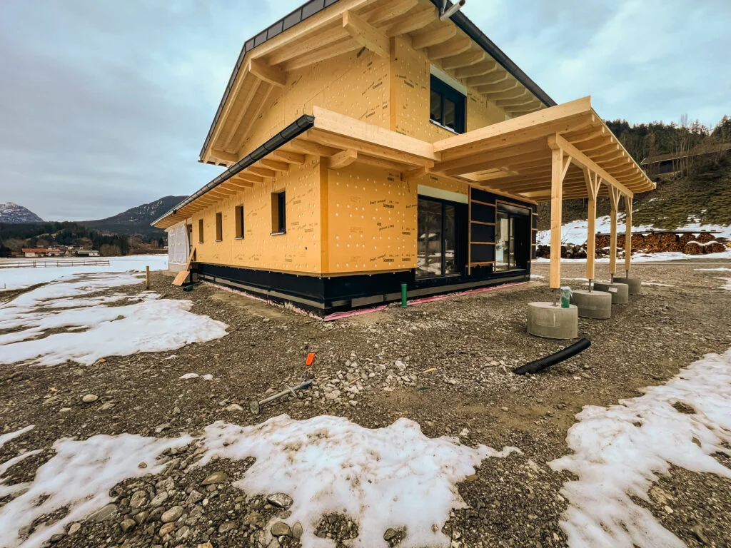 Zweistöckiges Holzhaus mit angebautem offenen Carport, sichtbare schwarze Abdichtungsbahn am Sockel; Schneereste, Bergkulisse