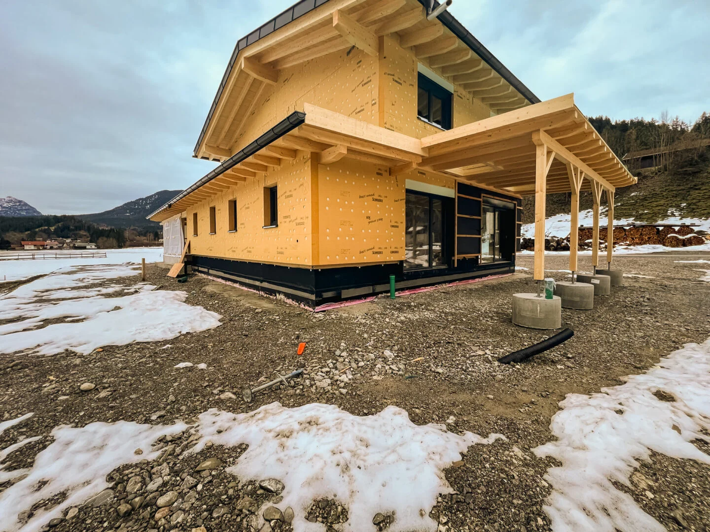 Zweistöckiges Holzhaus mit angebautem offenen Carport, sichtbare schwarze Abdichtungsbahn am Sockel; Schneereste, Bergkulisse
