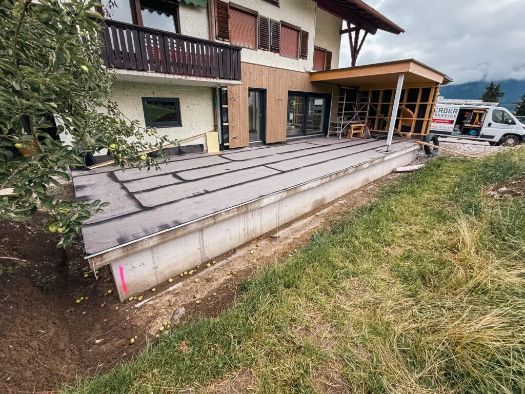 Haus mit Balkon und unvollendeter Terrasse, freigelegte Platten und Unterkonstruktion, Holzaufbau rechts, Rasen im Vordergrun