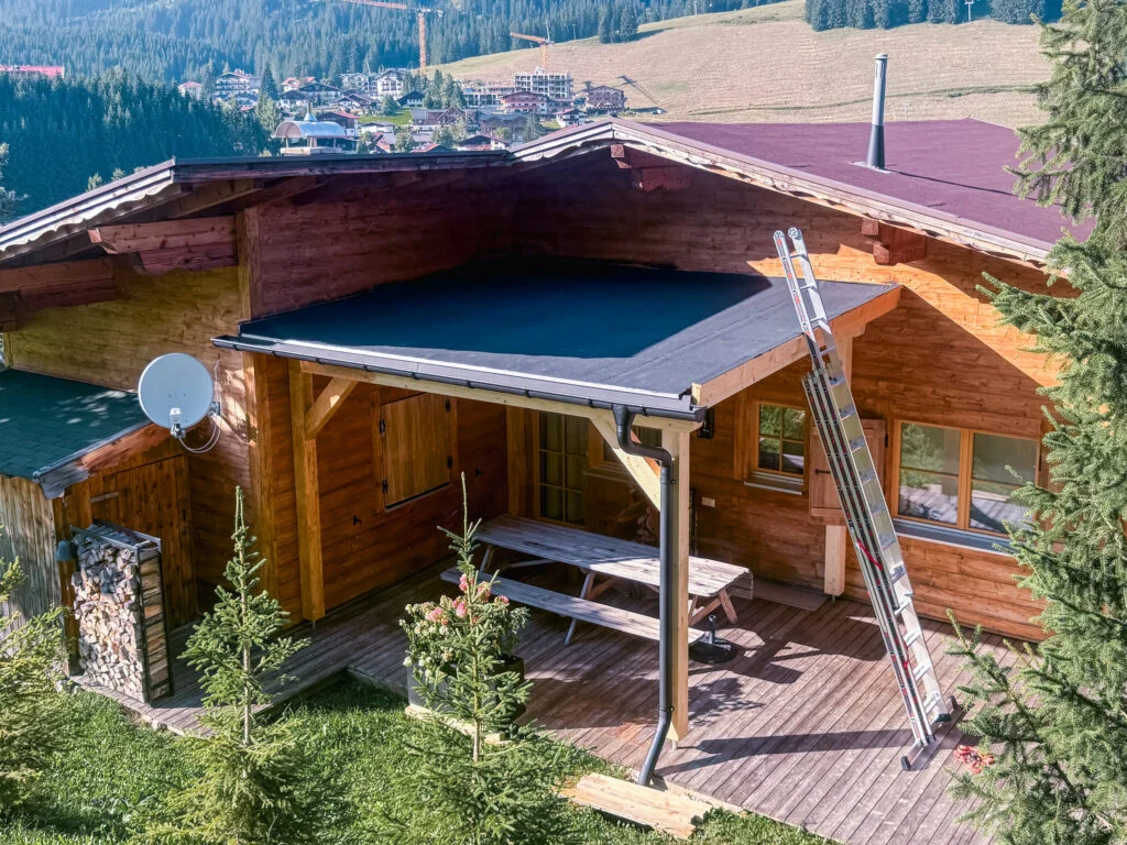 Holzhaus mit angebautem Carport/überdachtem Sitzplatz auf Holzdeck, dunkles Flachdach, Sitzbank, Satellitenschüssel, Bergblic