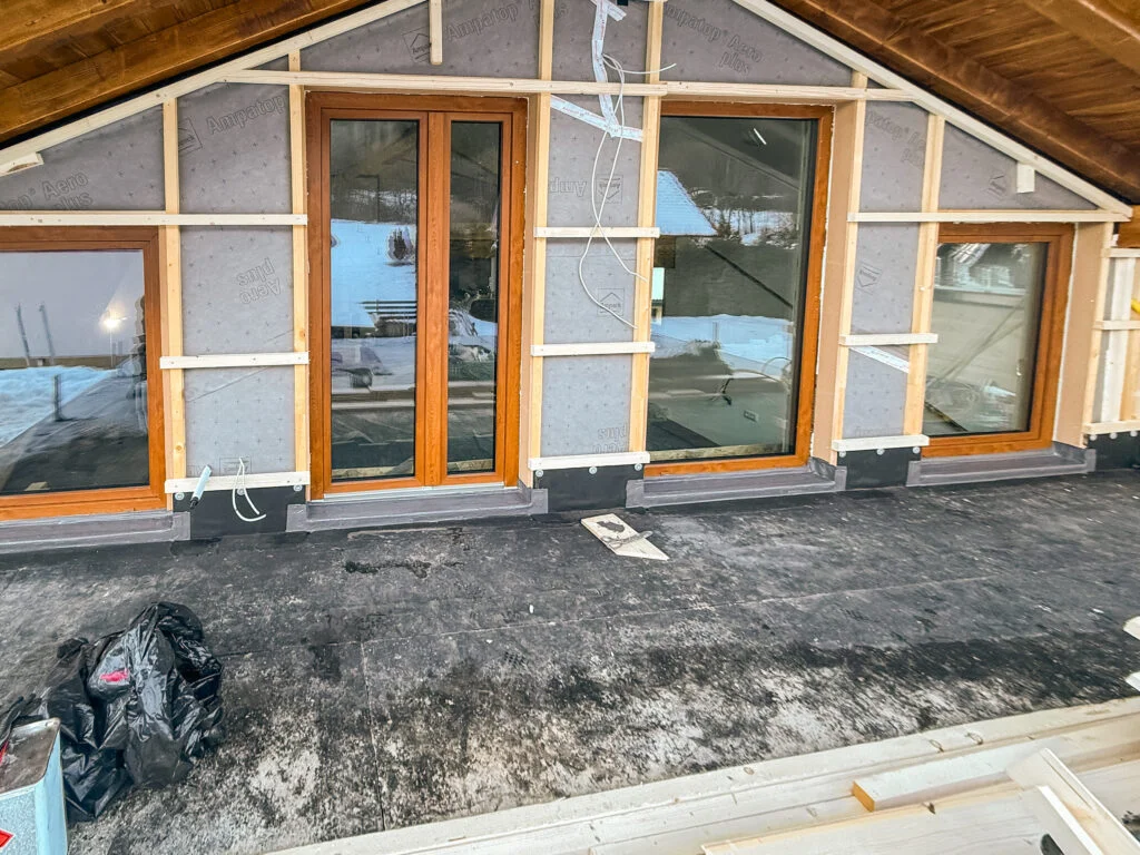 Dreiflügelige Holzfenstertüren an Fassade; Terrasse mit schwarzer Abdichtungsbahn, Arbeitsmaterialien und unvollendete Sockel