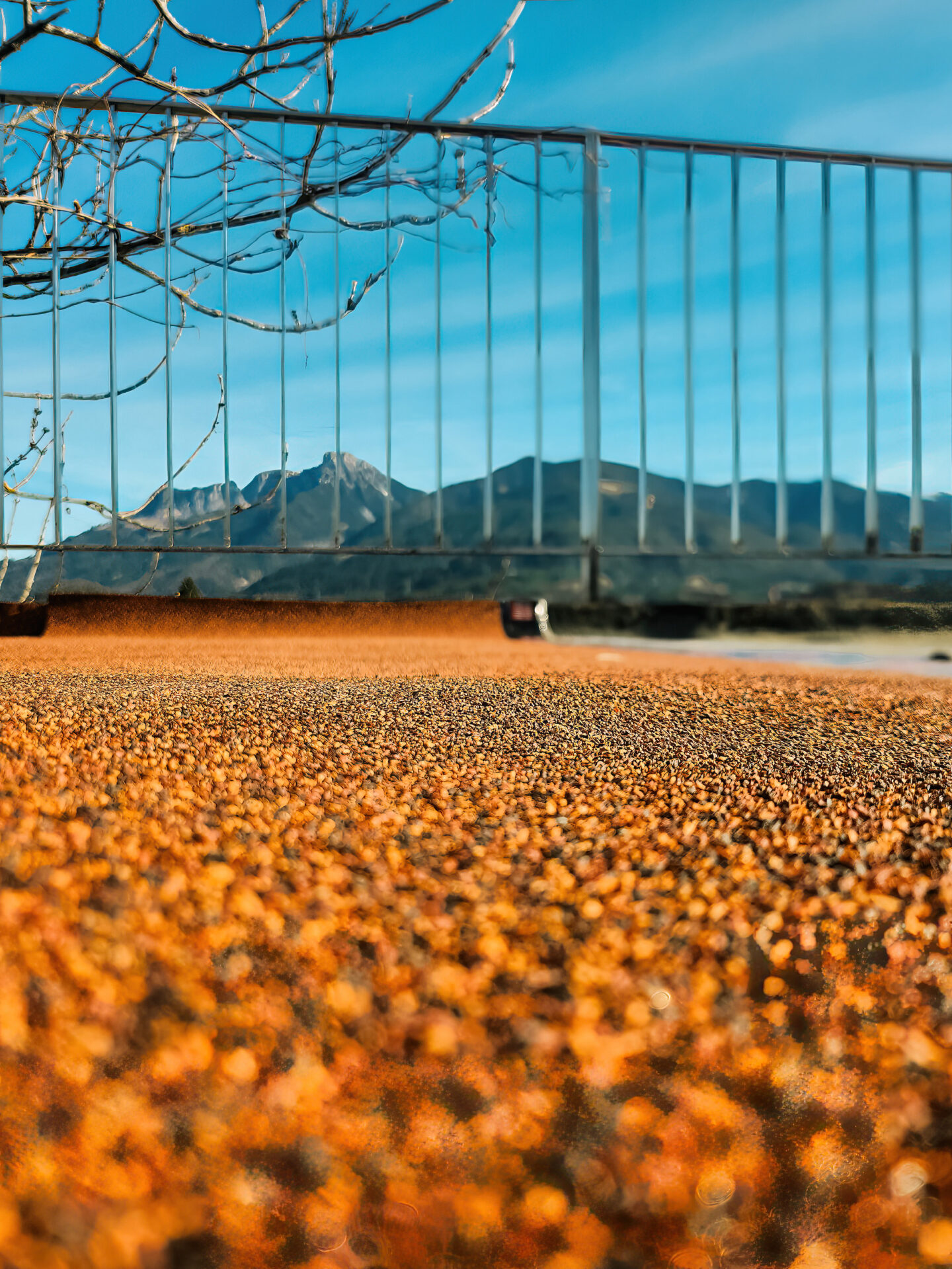 Orange körnige Terrassenoberfläche im Vordergrund, schmales Metallgeländer mittig, Alpenberge und blauer Himmel im Hintergrun