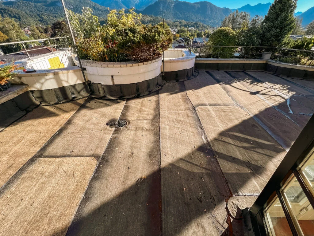 Flachdach mit aufliegenden Abdichtungsbahnen, niedriger Attika, Schatten eines Geländers und Bergkulisse im Hintergrund