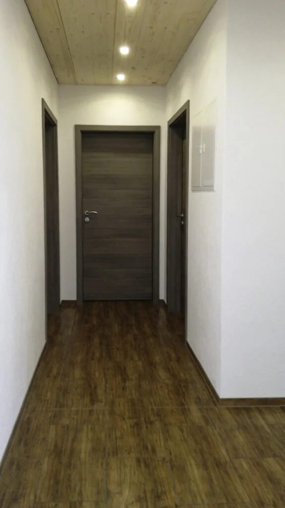 Schmaler wei&szlig;er Flur mit dunkler Wohnungst&uuml;r am Ende, dunklem Laminatboden und grauem Wandkasten rechts.