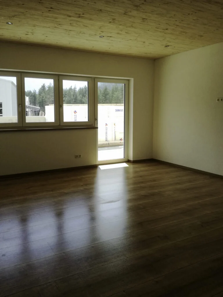 Helles, leeres Wohnzimmer mit Laminatboden, gro&szlig;e Fensterfront und Balkonzugang, wei&szlig;e W&auml;nde und Heizk&ouml;rper unterm Fenster.