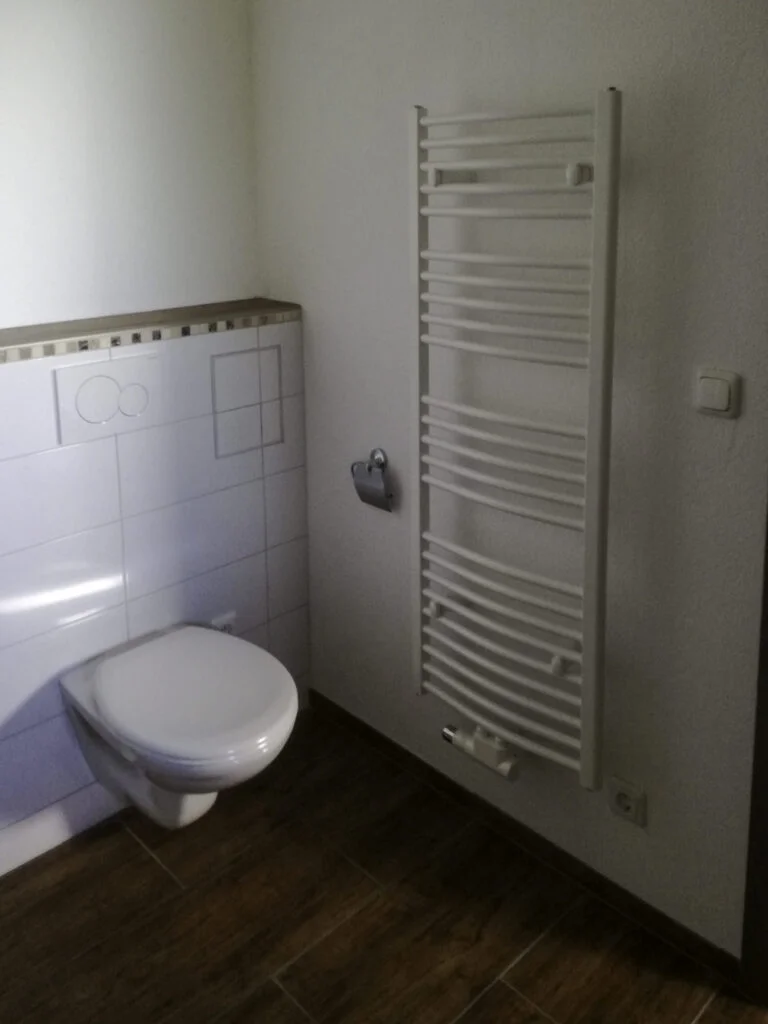 Wandh&auml;ngende Toilette, senkrechter wei&szlig;er Handtuchheizk&ouml;rper, Toilettenpapierhalter, dunkler Boden.