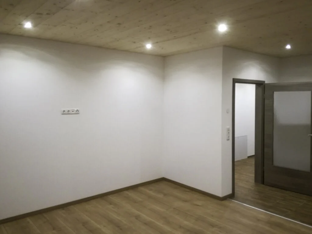 Leerstehender Mietwohnungsraum mit hellem Laminatboden, wei&szlig;en W&auml;nden, Deckenlampen und ge&ouml;ffneter T&uuml;r zum Nebenraum