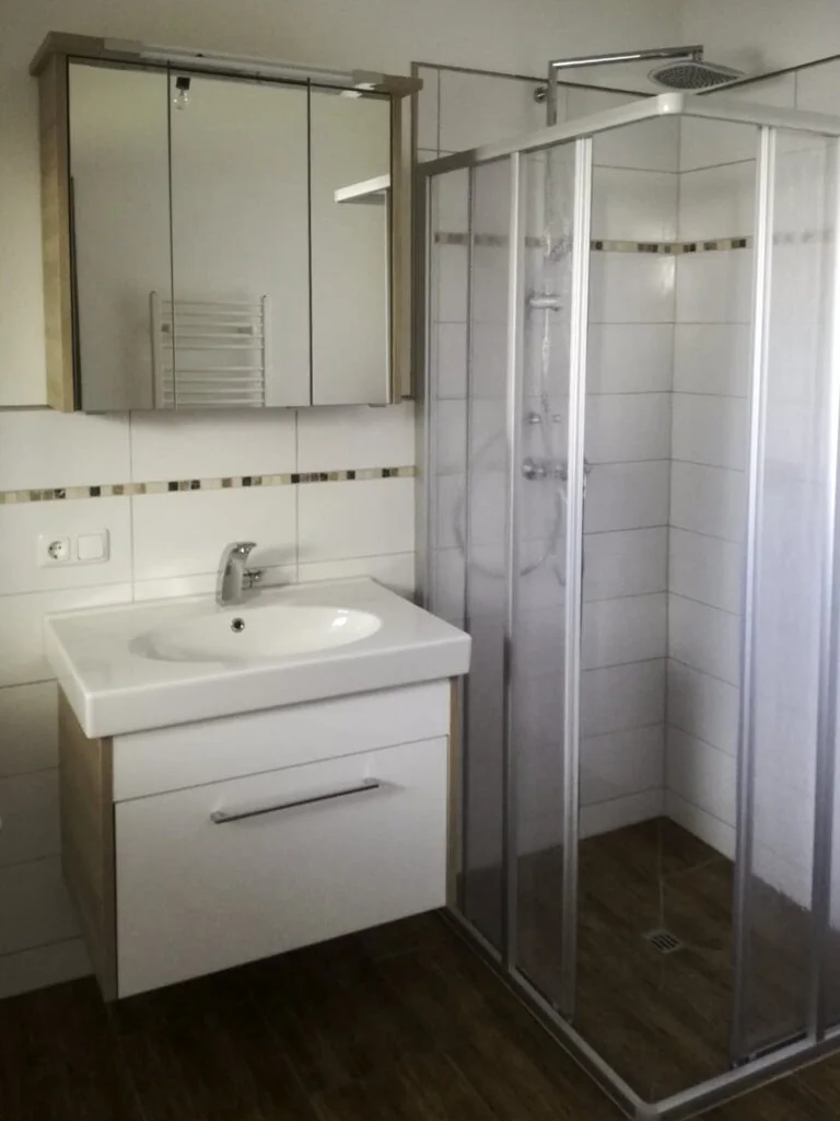 Badezimmer einer Mietwohnung in Stanzach: Waschbecken mit Unterschrank, Spiegel, Glast&uuml;r-Dusche und Handtuchheizk&ouml;rper.