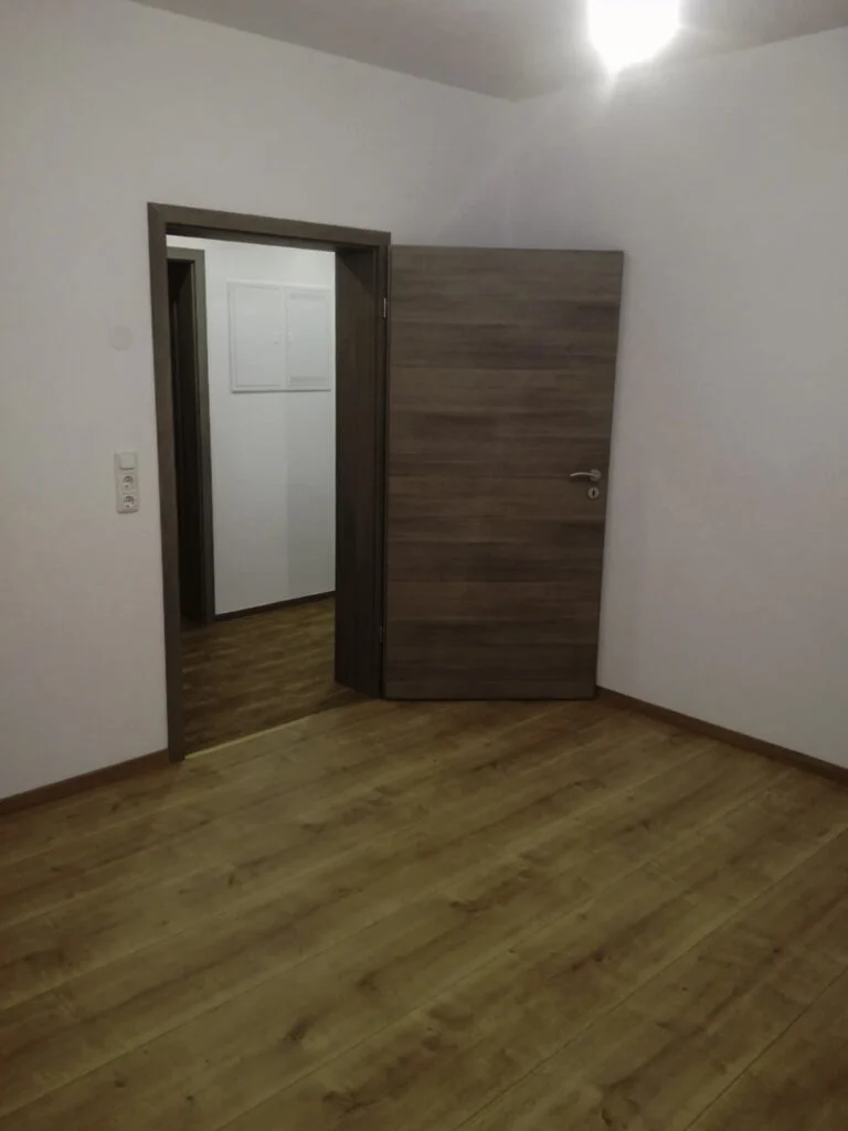 Leerer Raum in Wohnung mit Laminatboden, offene Holzinnent&uuml;r zum Flur, wei&szlig;e W&auml;nde, Lichtschalter sichtbar.