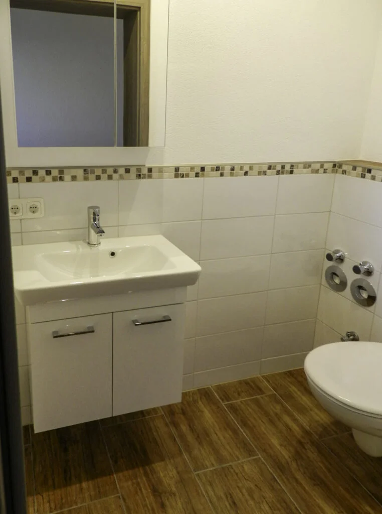 Badezimmer in Mietwohnung, wei&szlig;er Waschtisch mit Unterschrank, Mosaikbord&uuml;re an Wandfliesen, WC, holzfarbener Boden