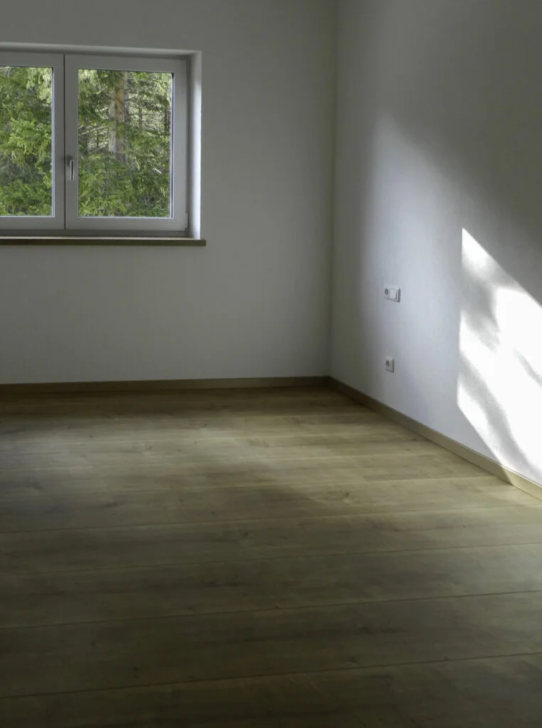 Leere Raumecke mit hellem Parkett, wei&szlig;en W&auml;nden, Fenster mit Gr&uuml;nblick und Sonnenschein.