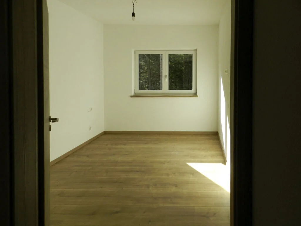 Leeres Zimmer in Mietwohnung: wei&szlig;e W&auml;nde, Holzfu&szlig;boden, Fenster an der Stirnwand, Sonneneinfall von rechts.
