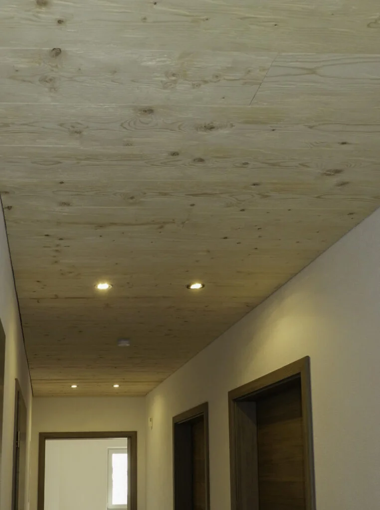 Flur einer Wohnanlage mit hellen W&auml;nden, Holzdecke, eingelassenen Deckenspots und mehreren T&uuml;ren rechts