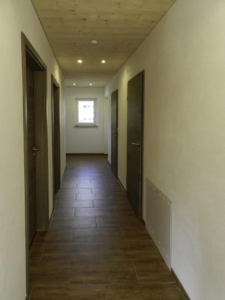 Schmaler Wohnungsflur mit dunklem Laminatboden, mehreren T&uuml;ren beidseitig, hellem Fenster am Ende und Deckenbeleuchtung.