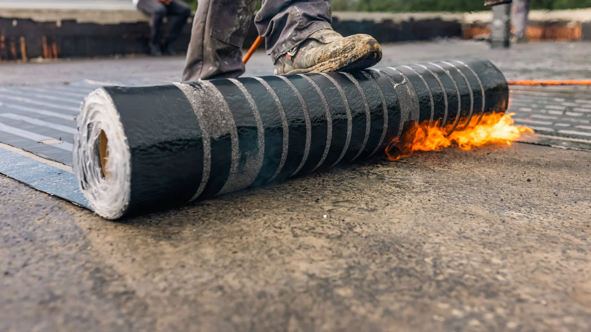 Stiefel auf Bitumenrolle; Propanbrenner erhitzt Naht der Dachabdichtung, Flamme am Bahnenrand.