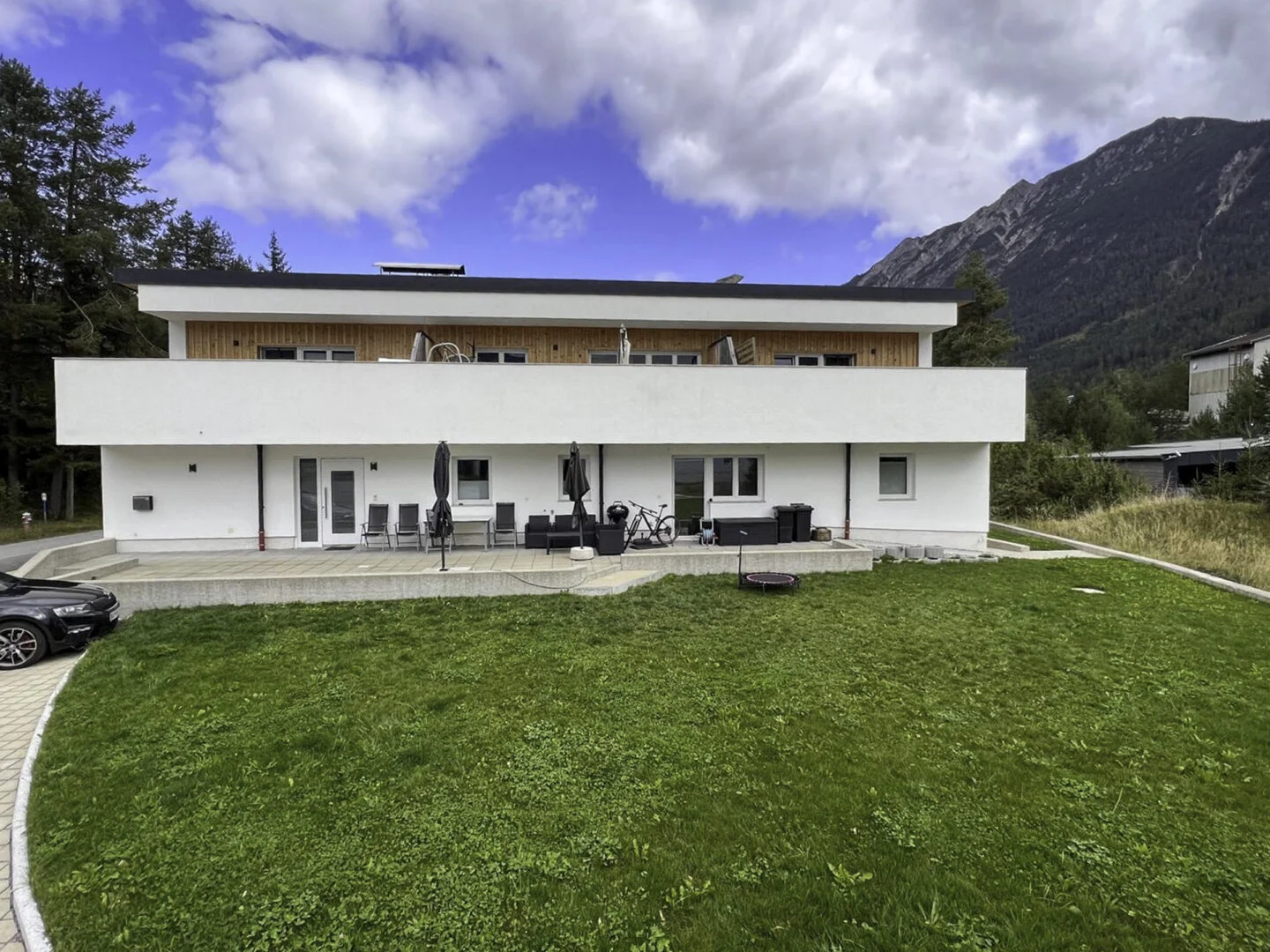 Weißes mehrstöckiges Wohngebäude mit Balkonen, Terrasse und Rasen, Berge im Hintergrund, bewölkter Himmel, Stanzach