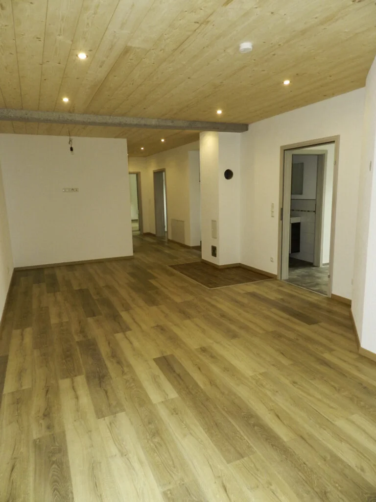 Heller leerer Wohnraum einer Mietwohnung mit Holzfu&szlig;boden, wei&szlig;en W&auml;nden, Deckenstrahlern, Flurzugang und Balkont&uuml;r.