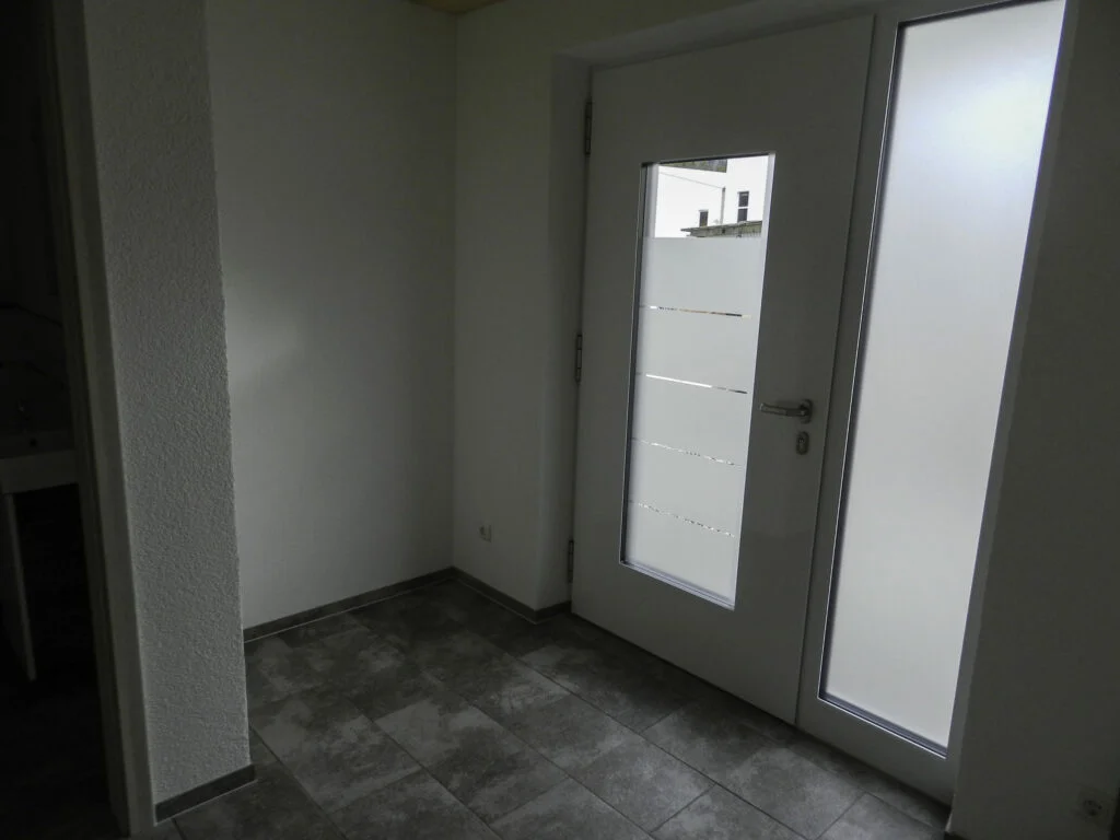 kleiner heller Innenraum mit wei&szlig; gestrichenen W&auml;nden, graue Bodenfliesen und eine seitliche T&uuml;r mit Milchglas-Einsatz