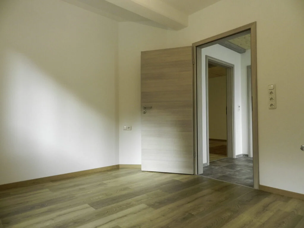 Leerer Raum in Mietwohnung (Stanzach) mit wei&szlig;en W&auml;nden, hellbraunem Holzfu&szlig;boden und offener Holzt&uuml;r zum Flur.